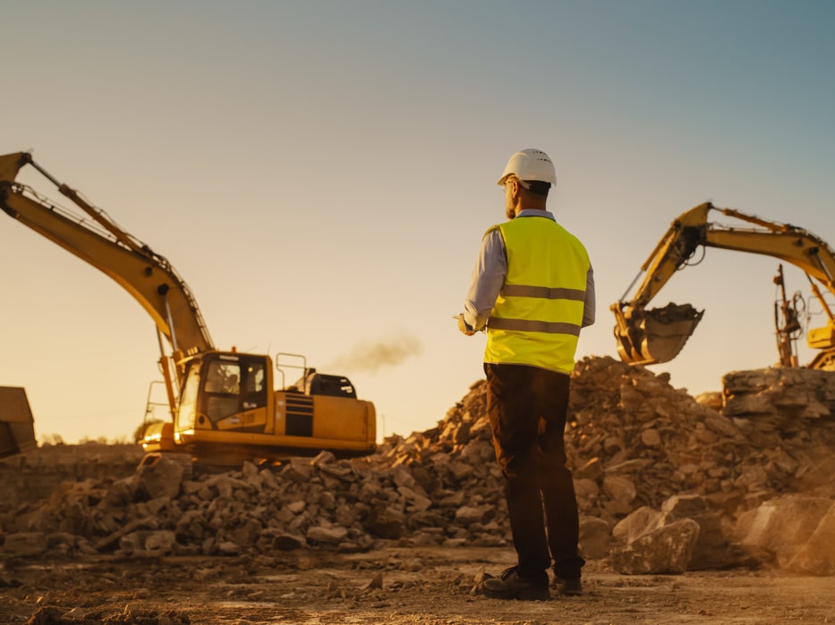 Caucasian Male Urban Planner Wearing Protective Goggles And Using Tablet On Construction Site On A Sunny Day. Man Inspecting Building Progress. Excavator Loading Materials Into Industrial Truck